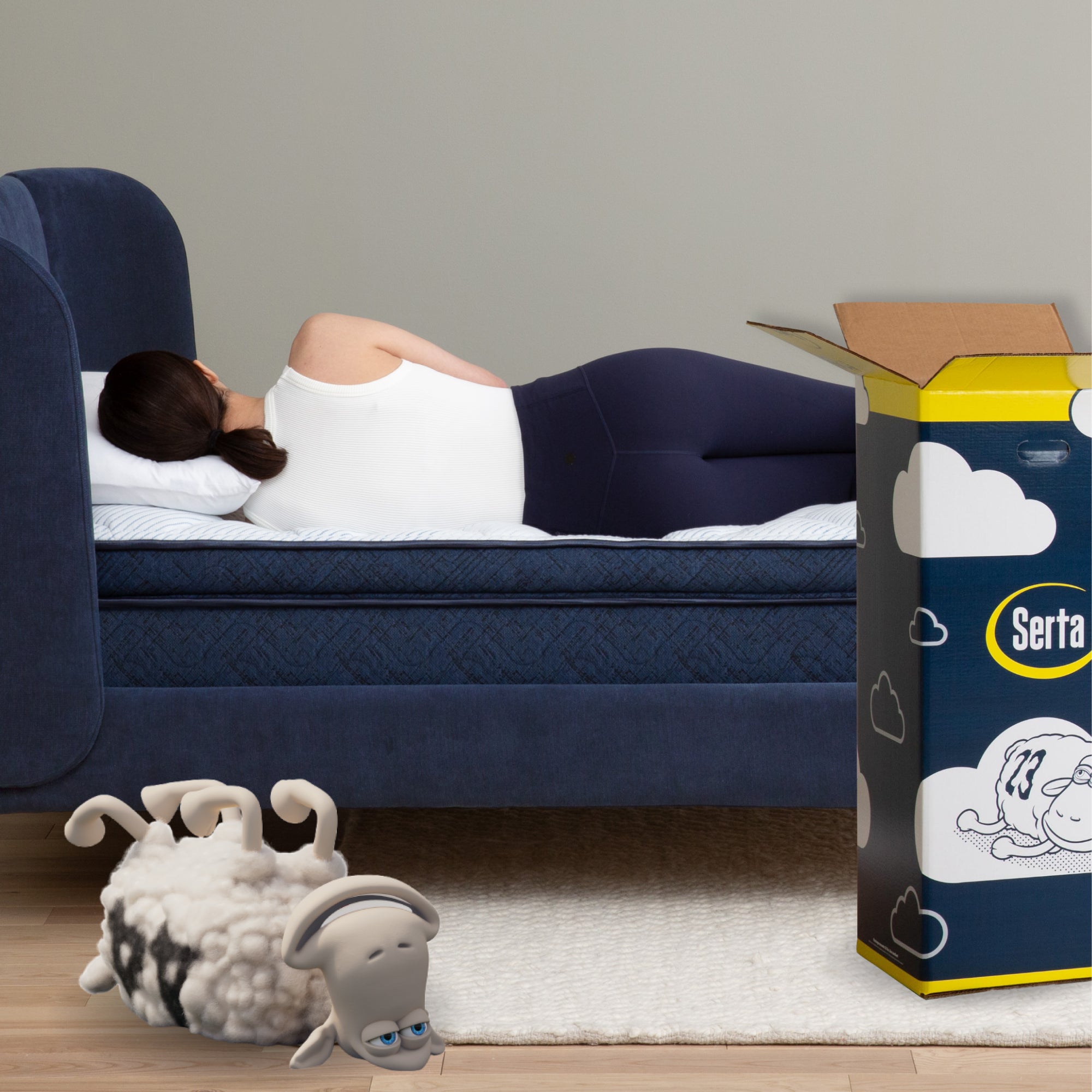 Woman laying on her left side of a Serta mattress in a box in a blue bed frame with her head on a pillow.  The packaging box is to the right of the bed at the foot with a Serta Sheep on the floor 