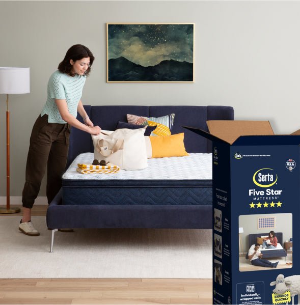 Woman standing to the left of a Serta Five Star mattress in a blue bedframe. with the box packaging sitting on the floor on the right.