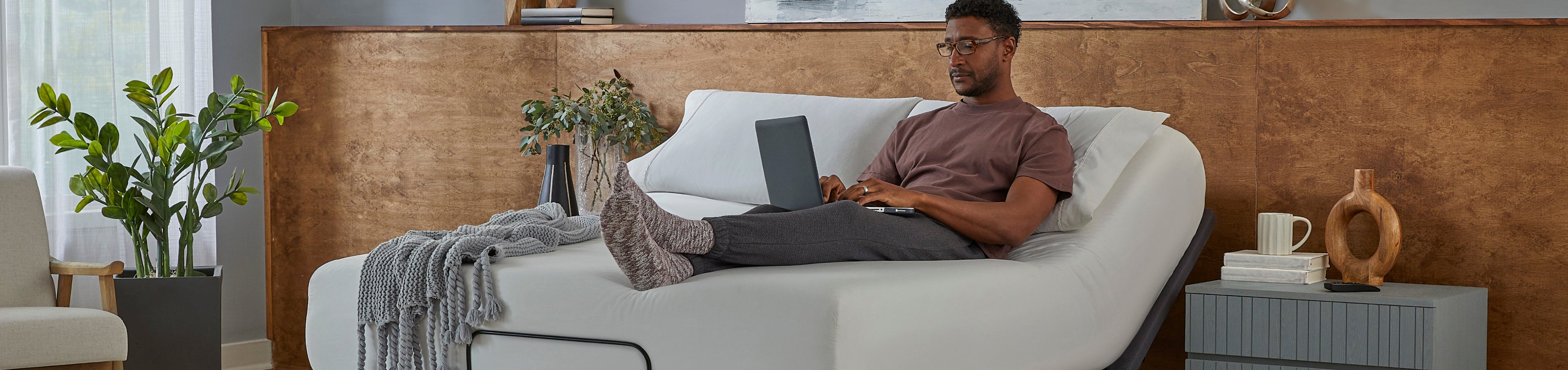 Man laying on a white mattress on a baselogic silver adjustable base working on his laptop
